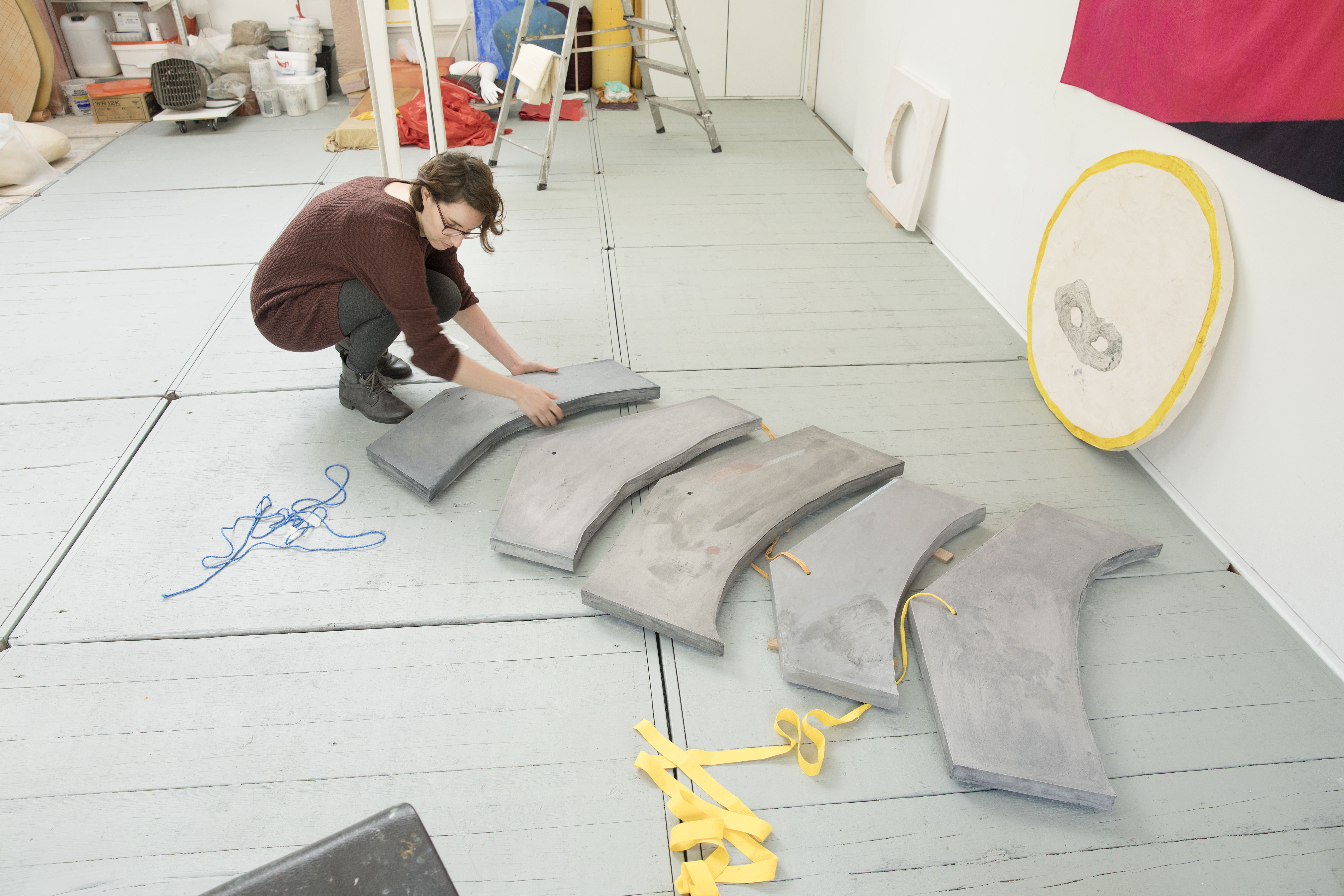 Eva Spierenburg werkt gehurkt op de vloer in haar atelier aan vijf grijze objecten